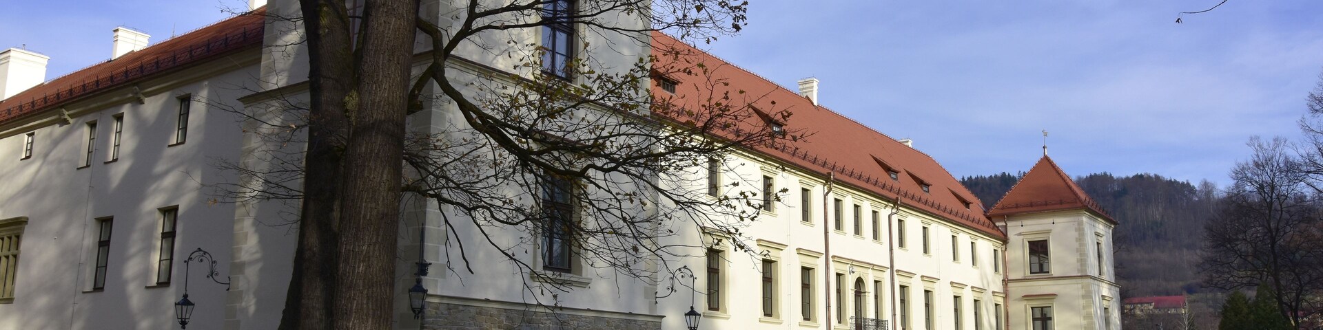 Um edifício grande e histórico com telhado de telhas vermelhas e paredes brancas, cercado por um gramado bem cuidado e uma árvore imponente.