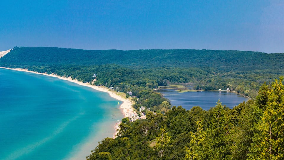 A coastal landscape with a sandy beach, a clear blue sea, and a forested hill in the background.