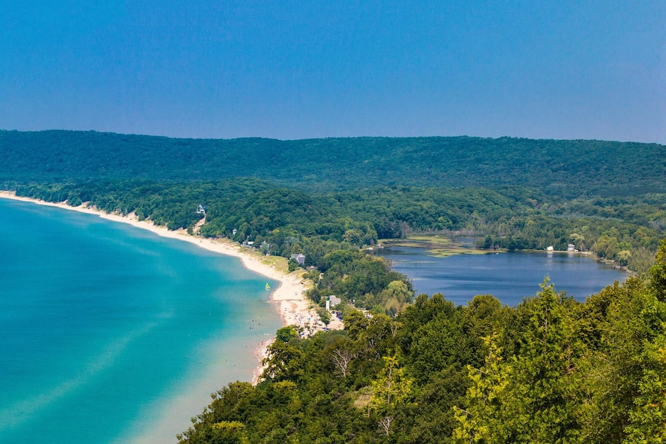 A coastal landscape with a sandy beach, a clear blue sea, and a forested hill in the background.