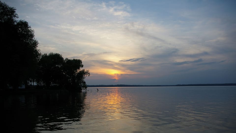 A sunset over a calm lake with trees on the left and a clear sky.