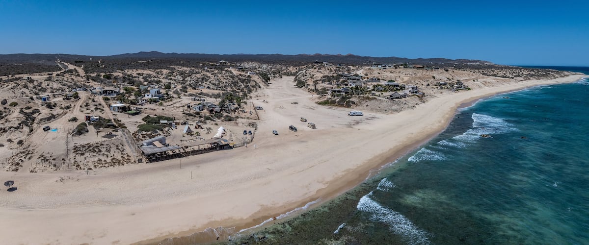 A coastal town with a beach, clear blue waters, and a hilly landscape in the background.
