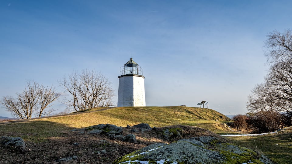 A lighthouse on a grassy hill with bare trees and rocks.