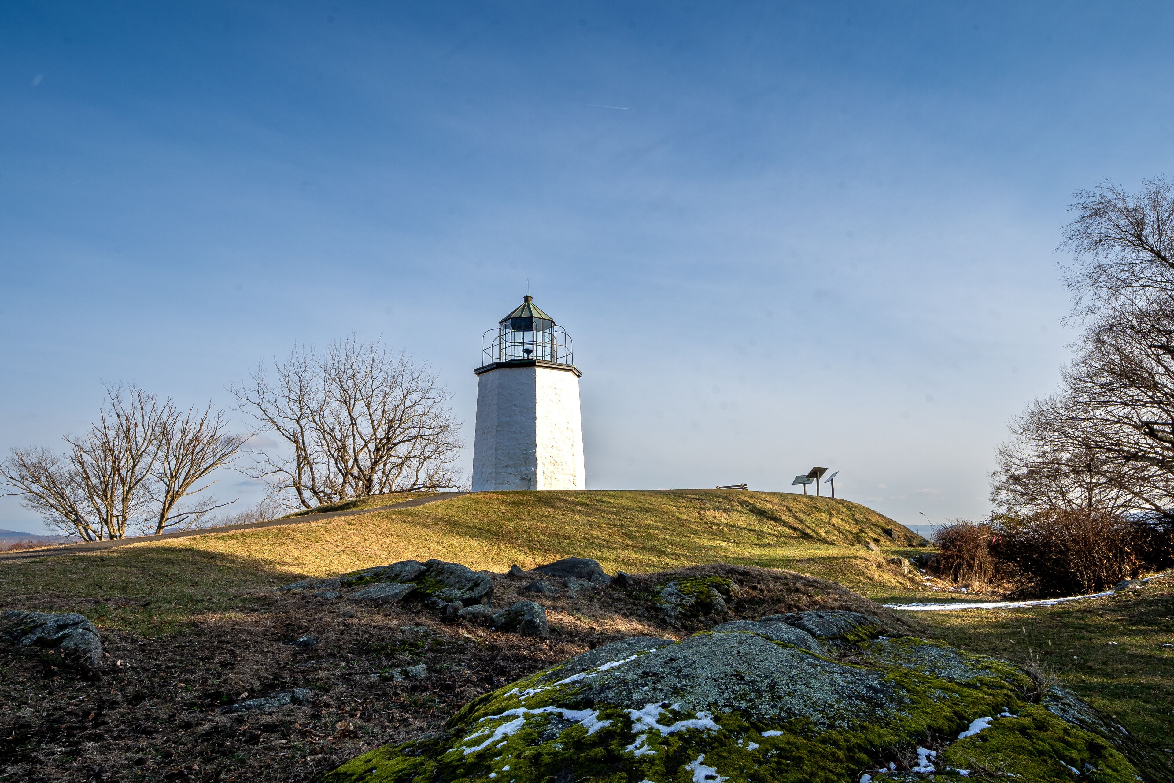 A lighthouse on a grassy hill with bare trees and rocks.