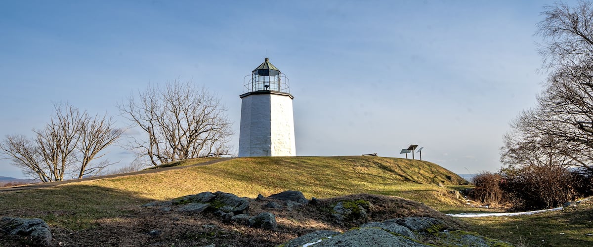 A lighthouse on a grassy hill with bare trees and rocks.