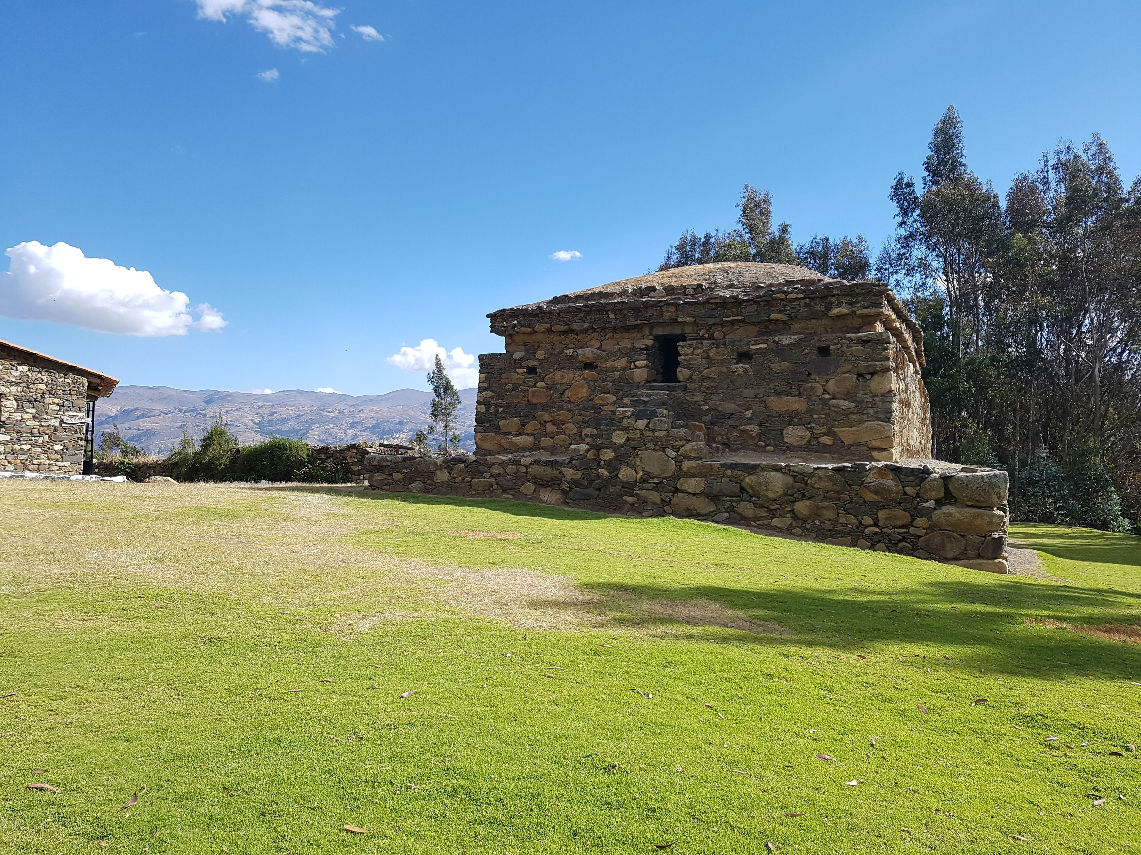 A stone building with a stone wall, a grassy field, and mountains in the background.