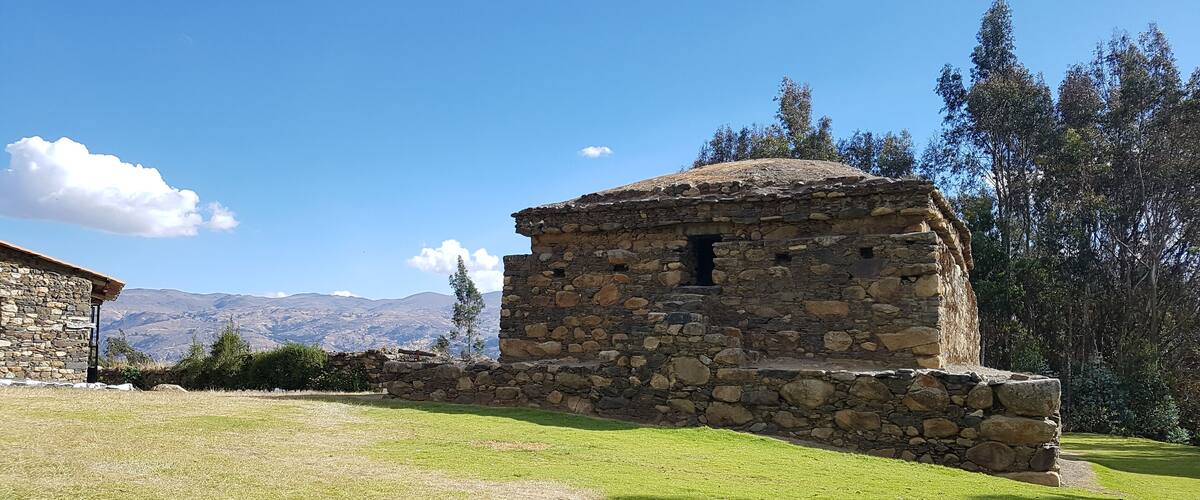 A stone building with a stone wall, a grassy field, and mountains in the background.