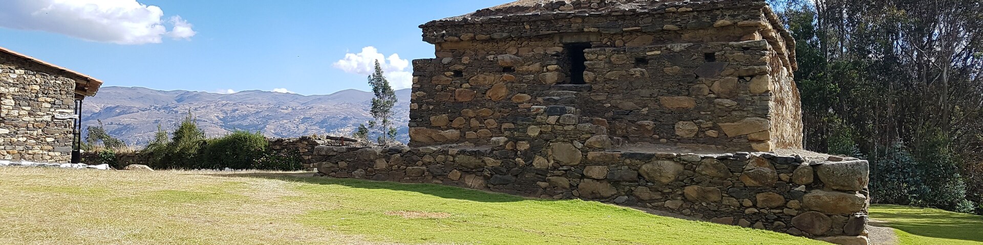 A stone building with a stone wall, a grassy field, and mountains in the background.