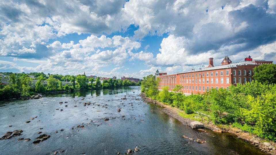 A river with rocks, a red brick building, and green trees under a cloudy sky.