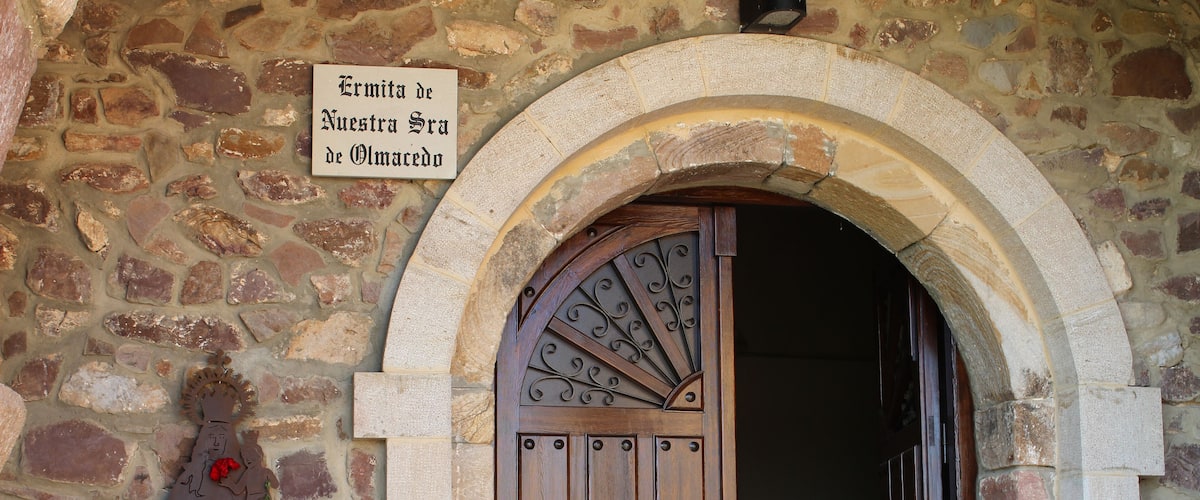A stone wall with a wooden door and a sign that reads "Ermitas de Nuestra Sra de Olmecada."