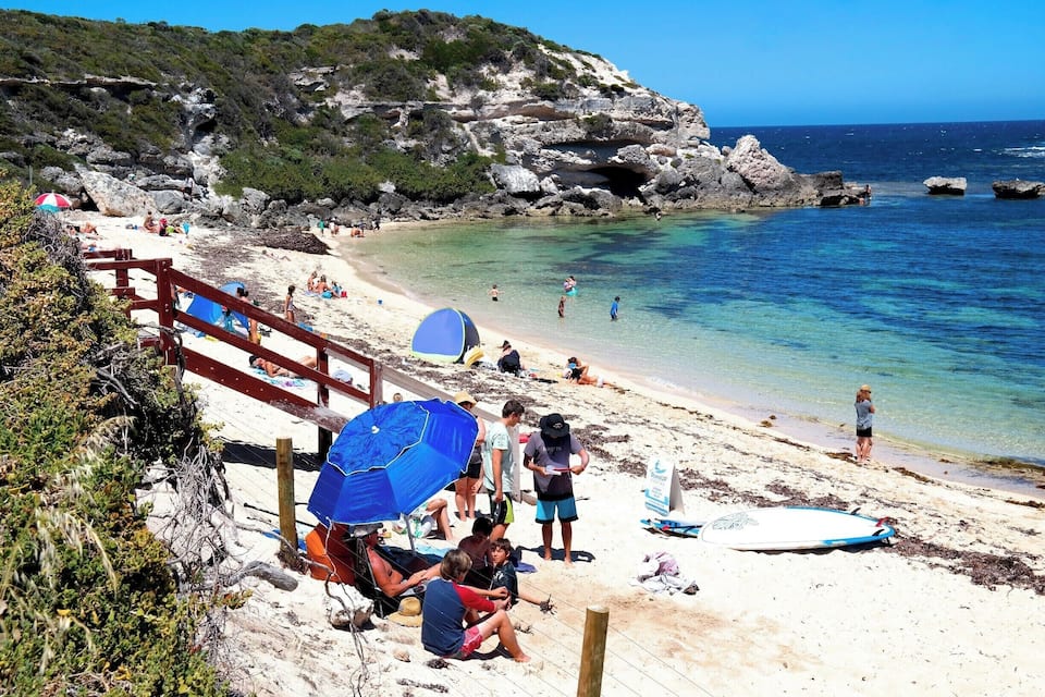 A beach with clear turquoise water, rocky cliffs, and a wooden walkway.