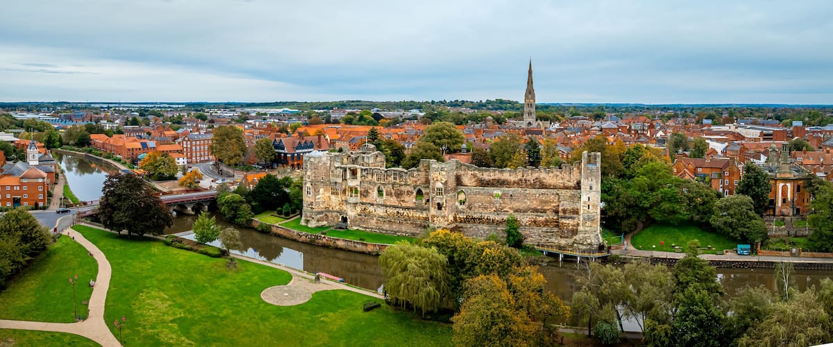 Een historisch kasteel met een rivier, omringd door een stad met rode daken en een kerktoren.