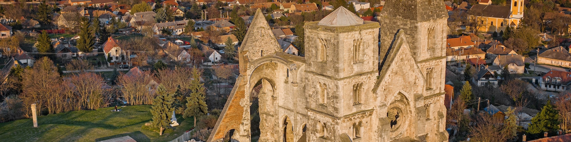 Vista aérea de una iglesia histórica con una torre destacada y arcos, rodeada por un pequeño pueblo y campos abiertos.