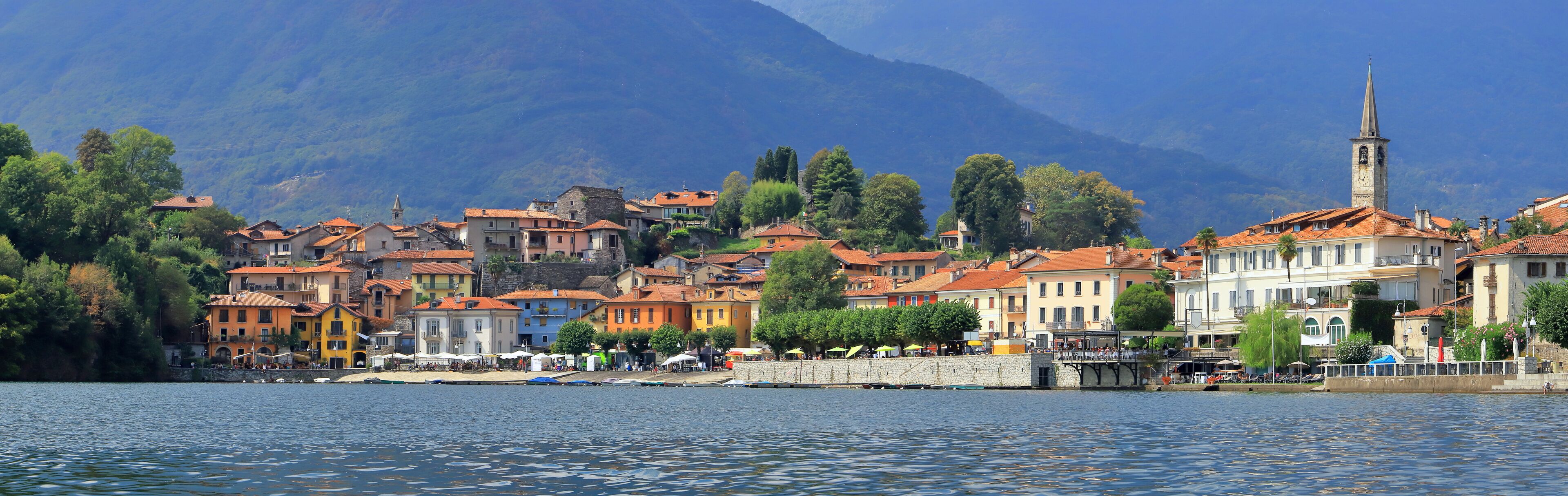 A lakeside town with colorful buildings, a church tower, and mountains in the background.