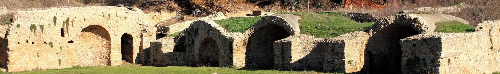 Ruinas antiguas de piedra con arcos y una colina cubierta de hierba al fondo.