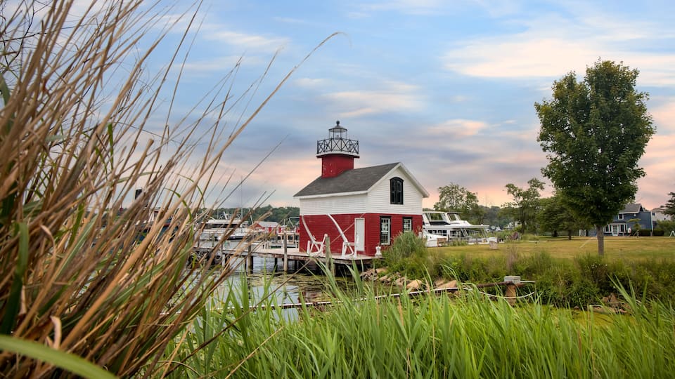 A red and white lighthouse by a dock with boats, surrounded by greenery and a cloudy sky.