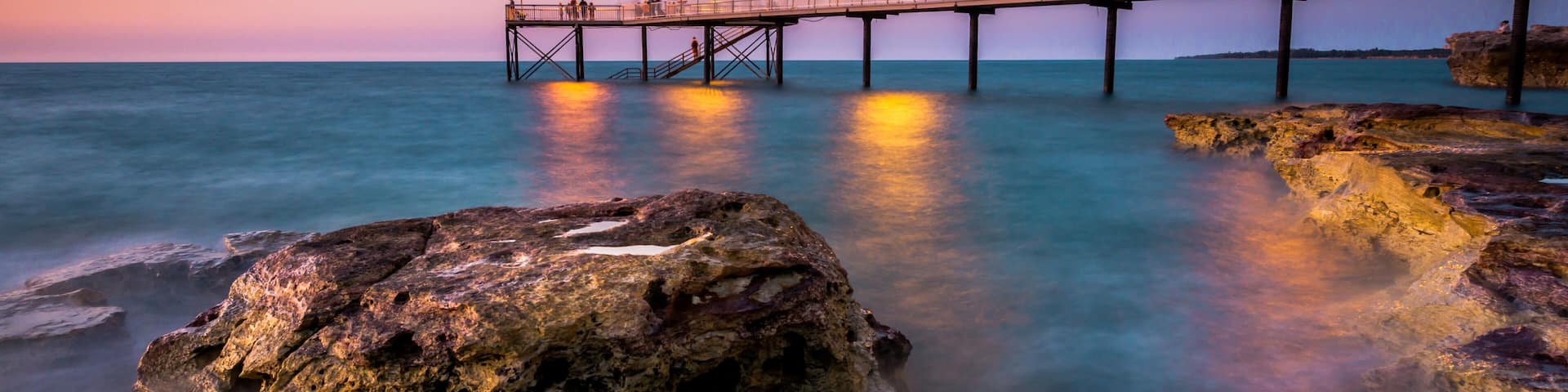 A pier extending into calm waters at dusk, with a rocky foreground and distant shoreline.
