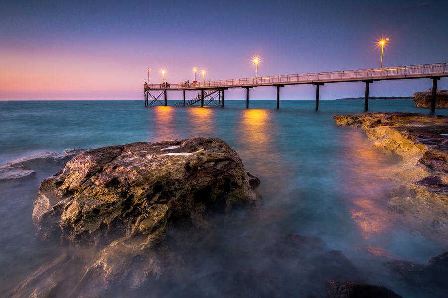 Un muelle se adentra en aguas tranquilas al atardecer, con un primer plano rocoso y una costa distante.