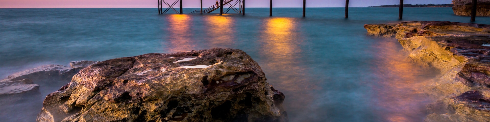 A pier extending into calm waters at dusk, with a rocky foreground and distant shoreline.