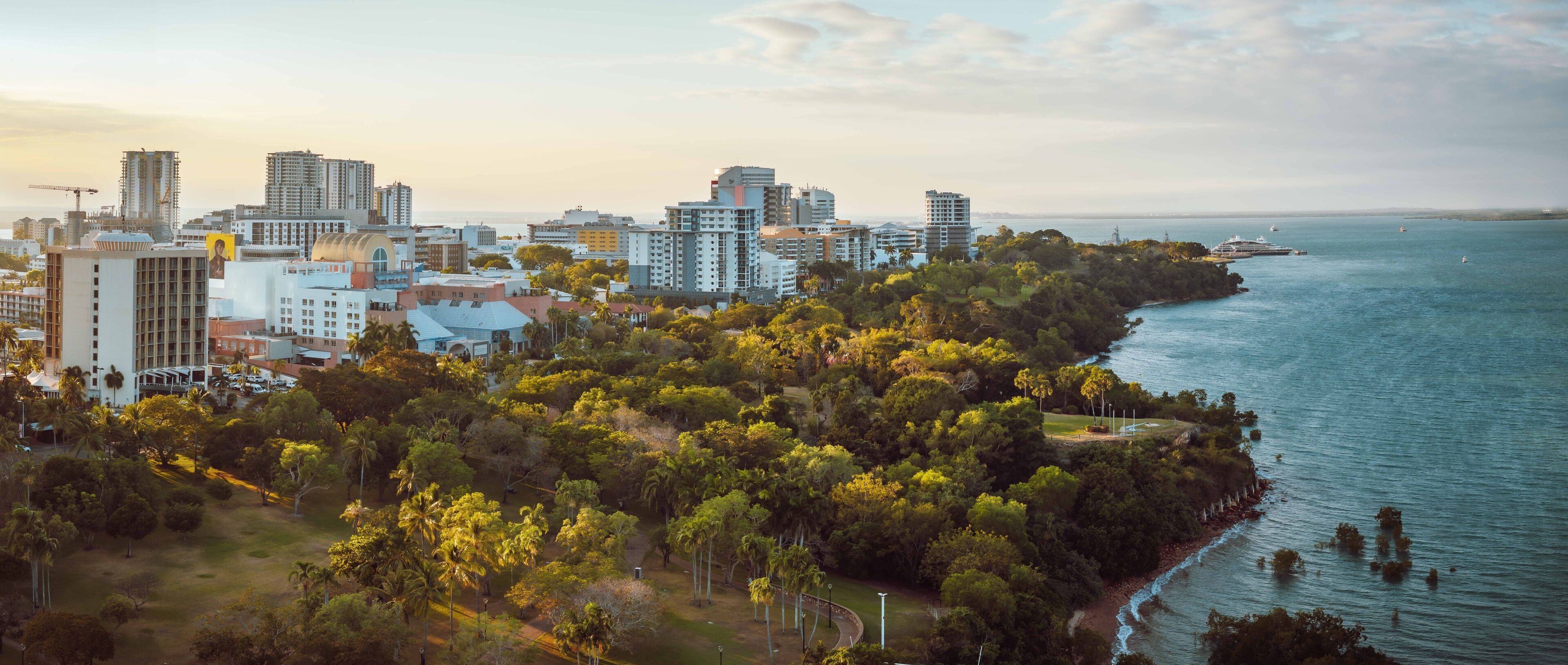 A coastal cityscape with dense greenery, high-rise buildings, and a calm sea.