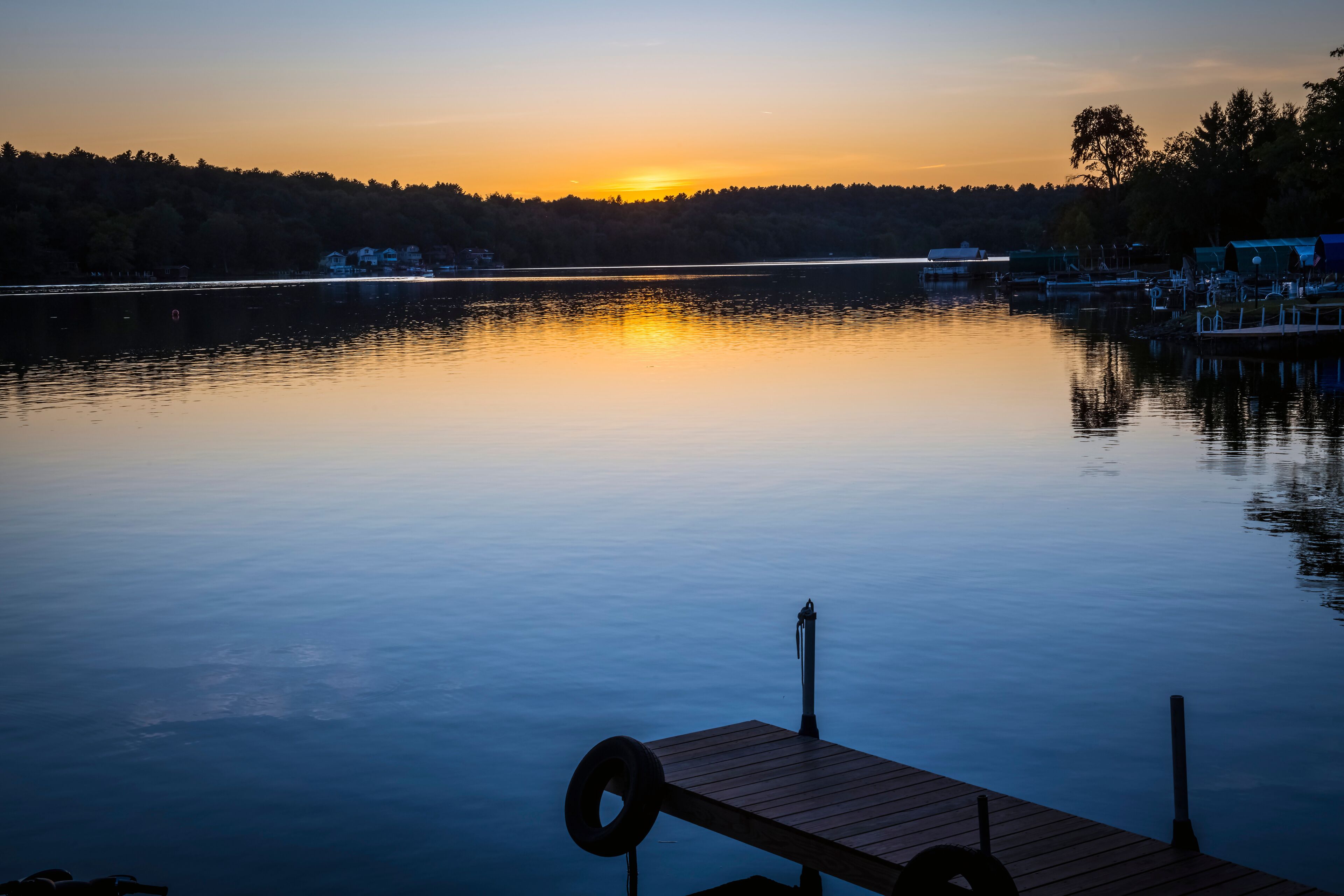 A calm lake at sunset with a wooden dock in the foreground and houses along the shore.