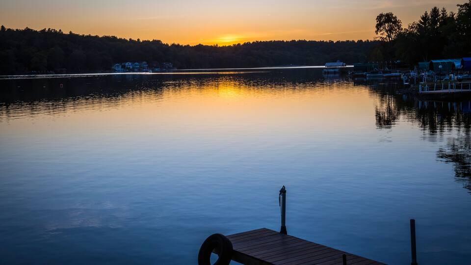 A calm lake at sunset with a wooden dock in the foreground and houses along the shore.