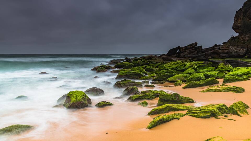 A beach with green moss-covered rocks and a sandy shore under a cloudy sky.