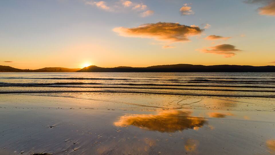 A beach at sunset with waves, a clear sky, and distant hills.