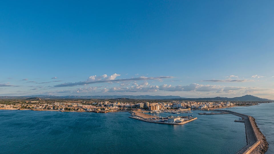 A coastal cityscape with a marina, a long pier, and a mountain range in the background.