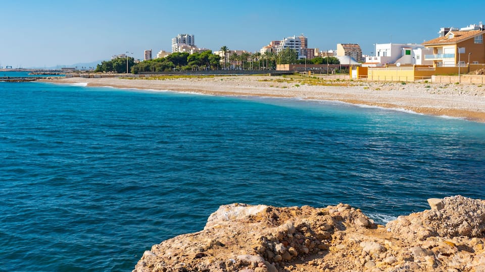 A coastal cityscape with a rocky shoreline, clear blue sea, and residential buildings.