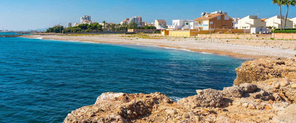A coastal cityscape with a rocky shoreline, clear blue sea, and residential buildings.