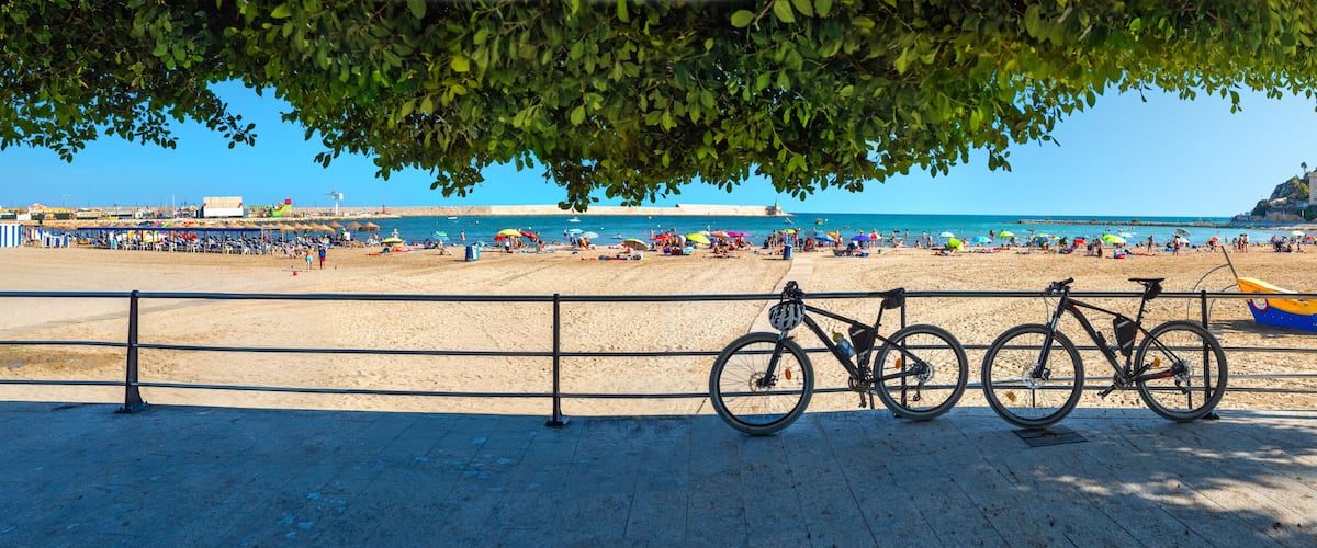 A beach with a bicycle parked by the railing, a sandy shore, and a clear blue sky.