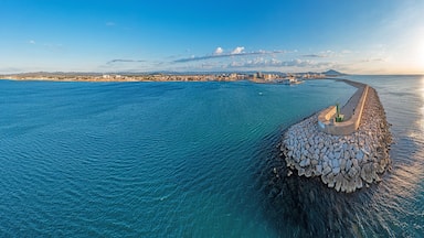 Un paysage urbain côtier avec une digue imposante qui s’avance dans la mer, un ciel dégagé et des montagnes lointaines.
