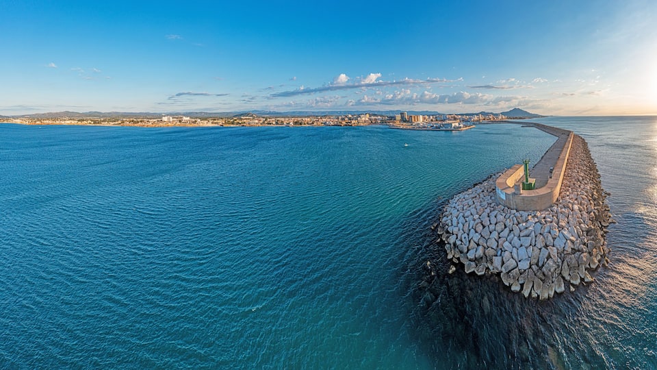 A coastal cityscape with a prominent breakwater extending into the sea, a clear sky, and distant mountains.