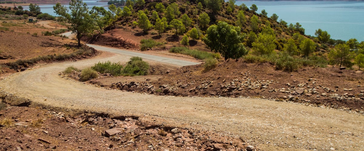 A winding dirt road through a hilly landscape with sparse trees and a lake in the background.