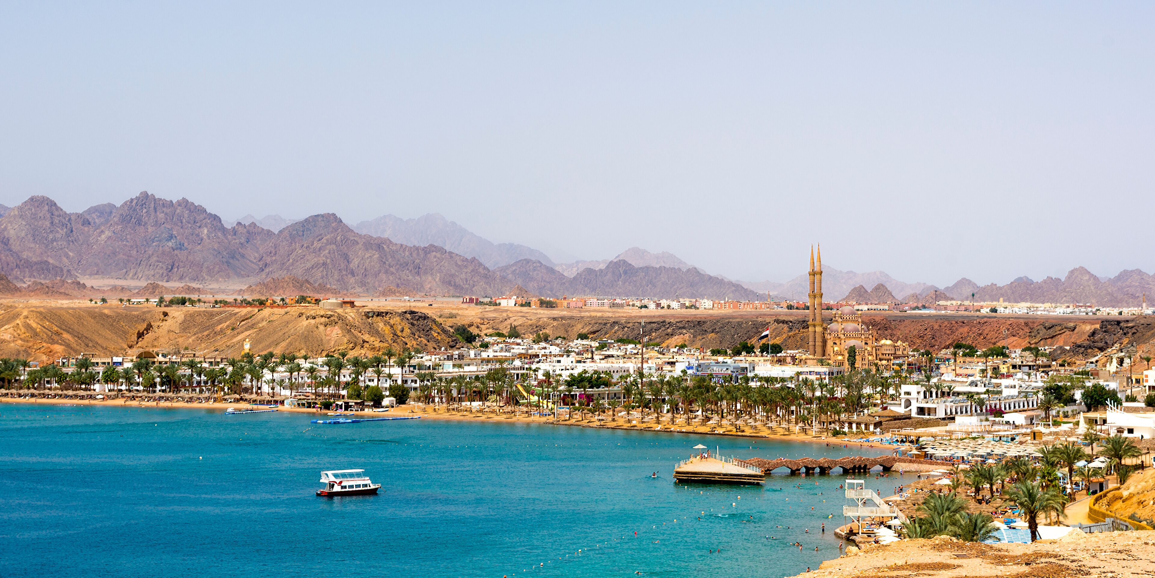 A coastal city with a prominent mosque, palm-lined beaches, and mountains in the background.