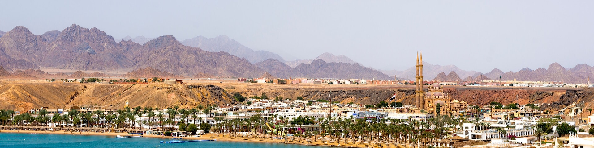 A coastal city with a prominent mosque, palm-lined beaches, and mountains in the background.
