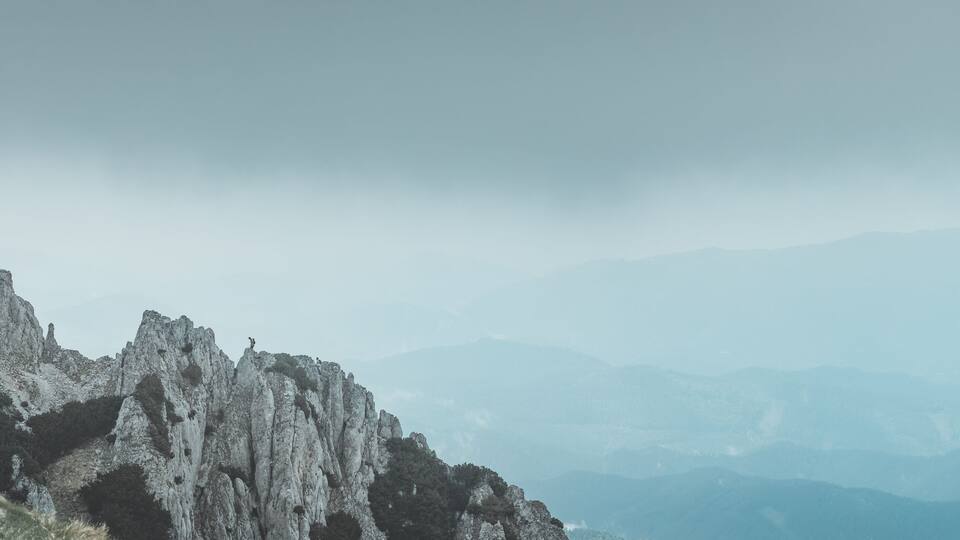 A rugged mountain landscape with a prominent rocky cliff and distant mountains under a cloudy sky.