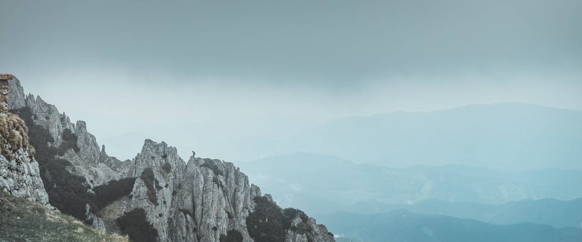A rugged mountain landscape with a prominent rocky cliff and distant mountains under a cloudy sky.
