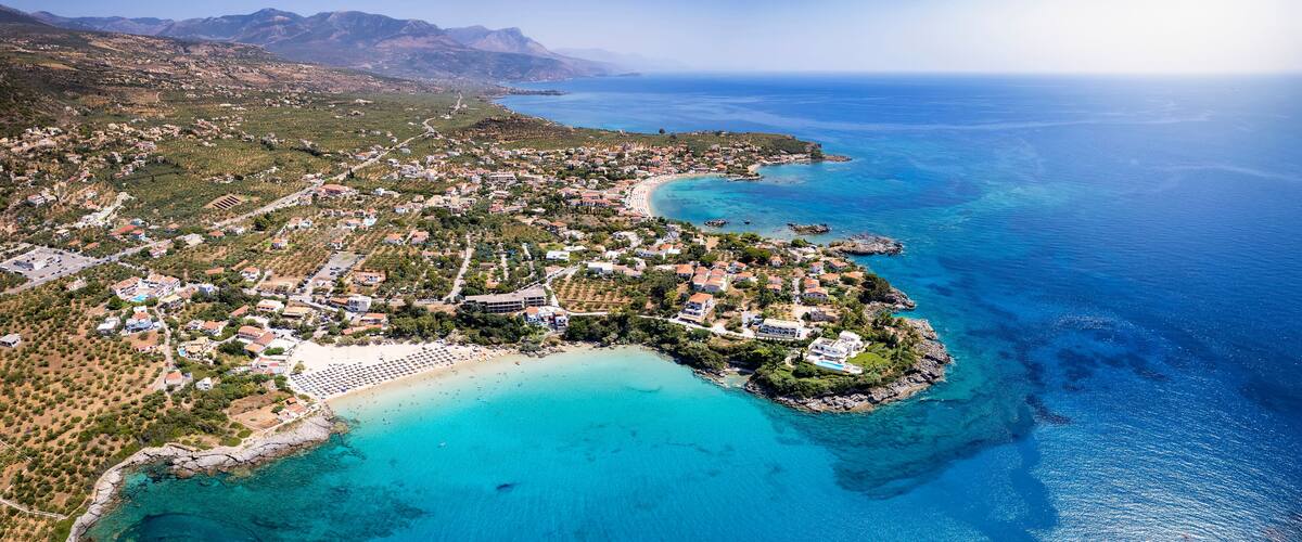 An aerial view of a coastal town with a beach, clear blue waters, and mountainous terrain in the background.
