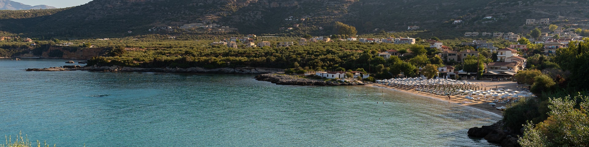 Uma cidade costeira com águas turquesas cristalinas, uma praia arenosa e montanhas ao fundo.