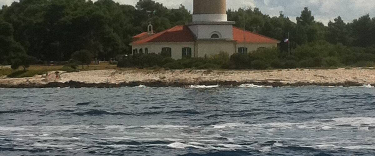 A lighthouse with a red roof, situated on a rocky shoreline with trees in the background.