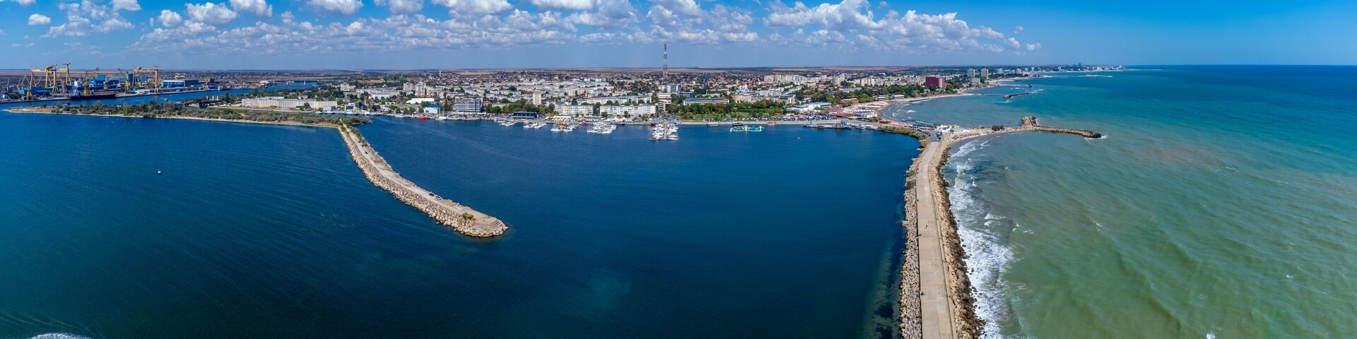 Una ciudad costera con un puerto deportivo, un rompeolas y grúas industriales.