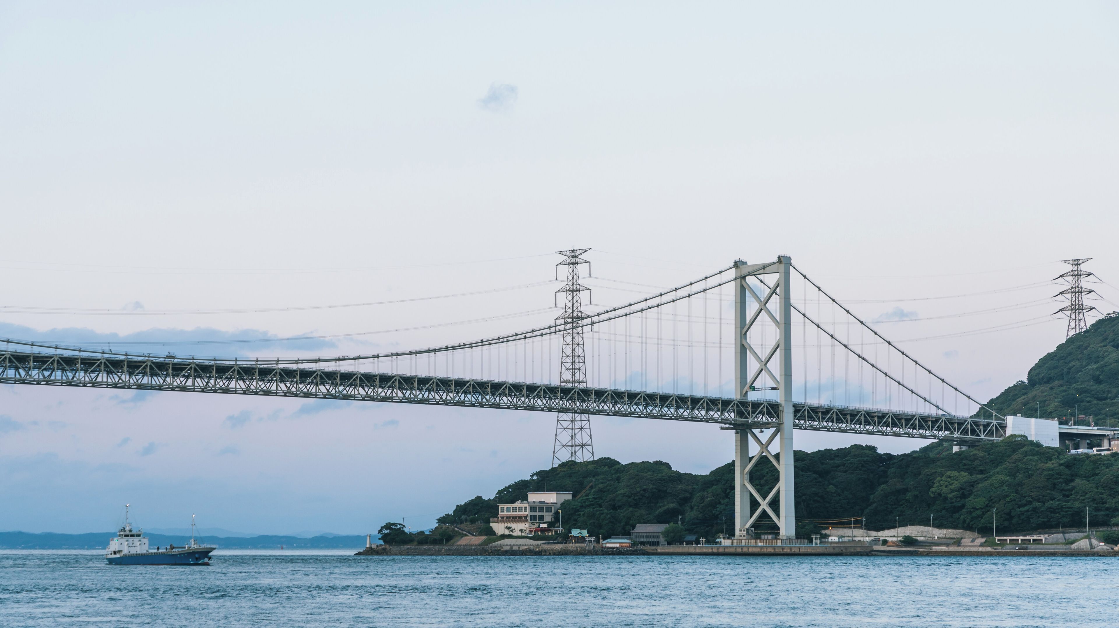 A suspension bridge over a body of water with a ship in the foreground and a hilly landscape in the background.