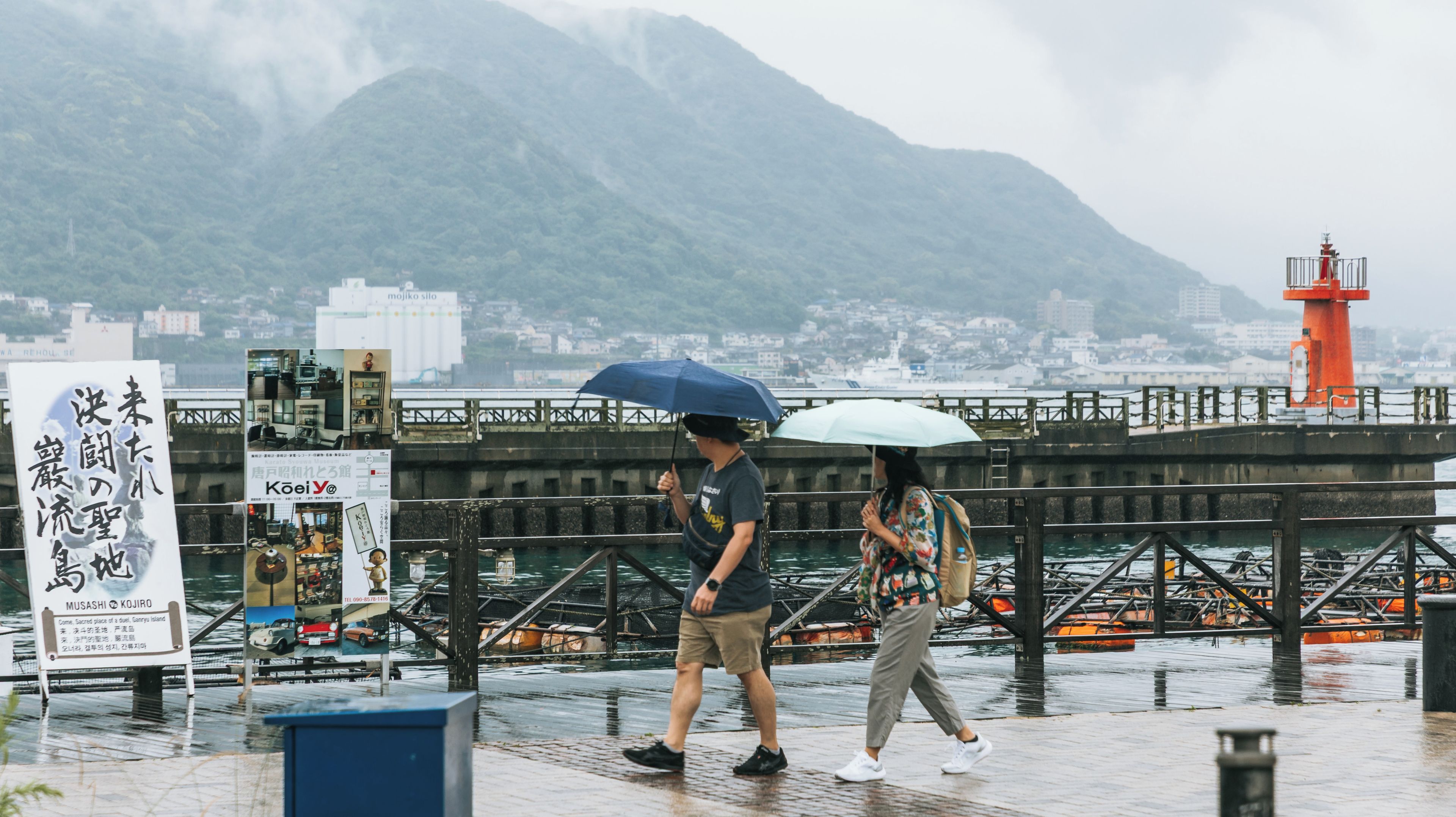 Um casal caminhando sobre uma ponte molhada, com paisagem urbana e montanhas ao fundo.