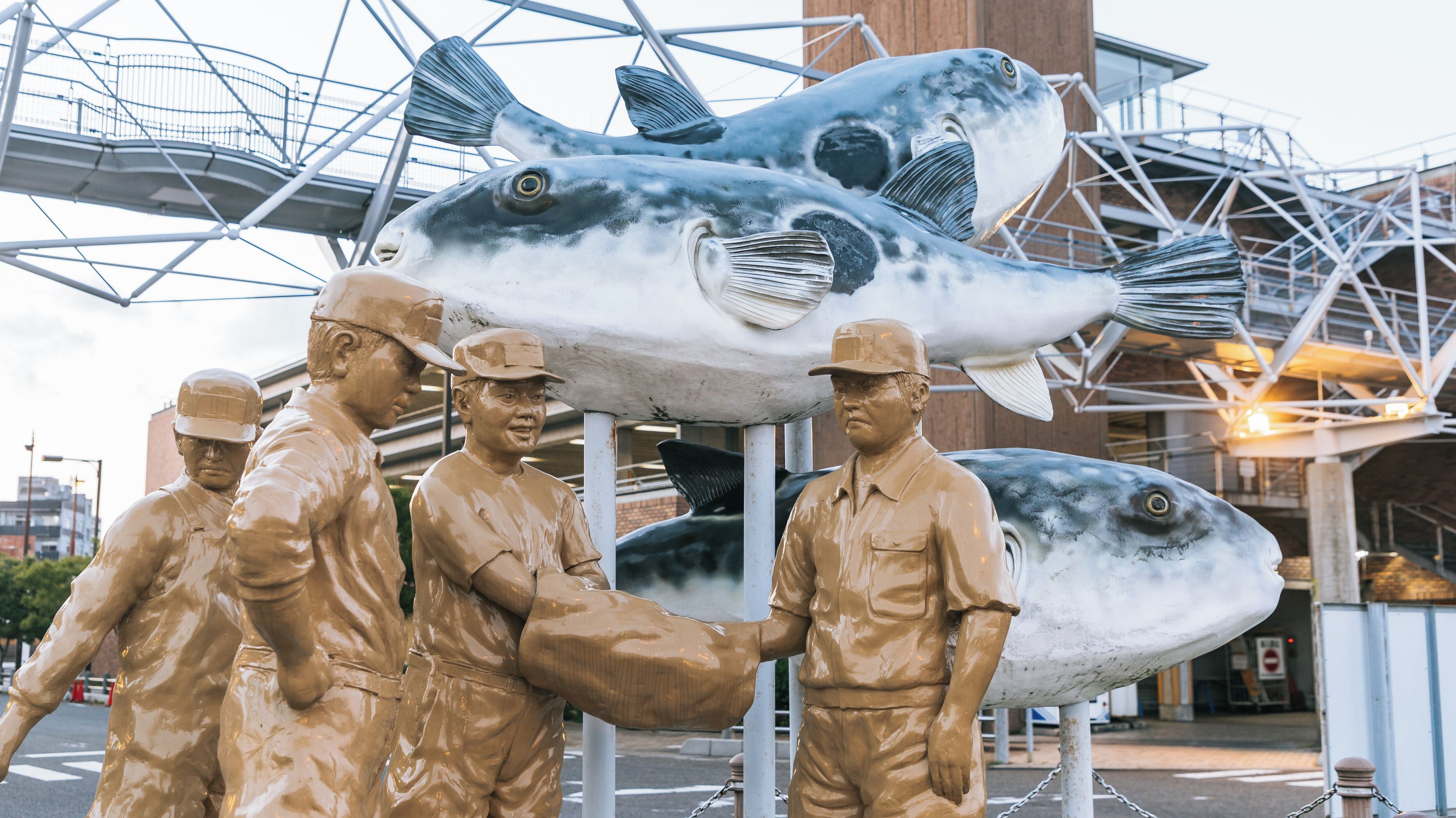 A large fish sculpture with four human figures in front of a metal structure.