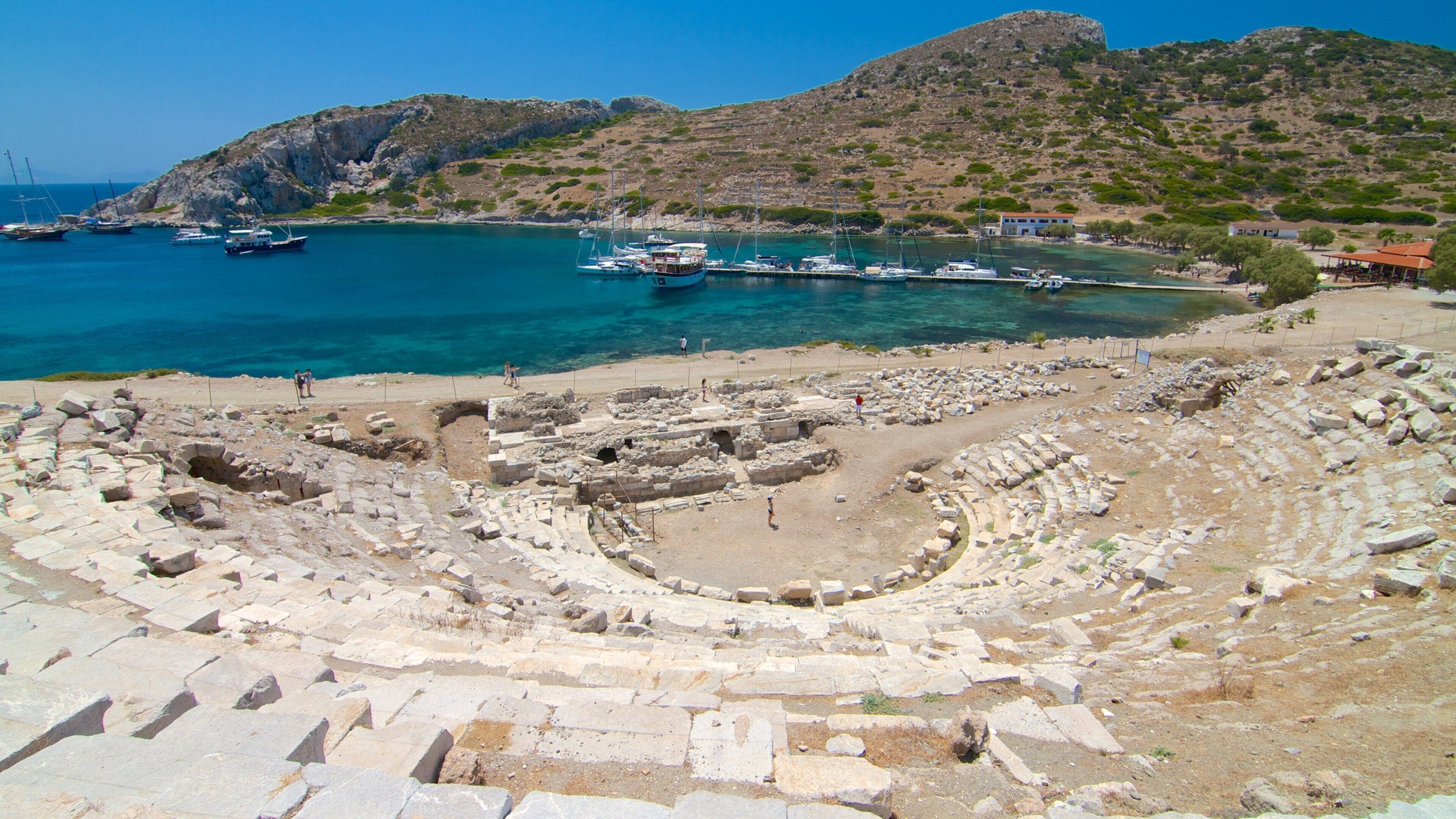 An ancient amphitheater with ruins and boats in the background.