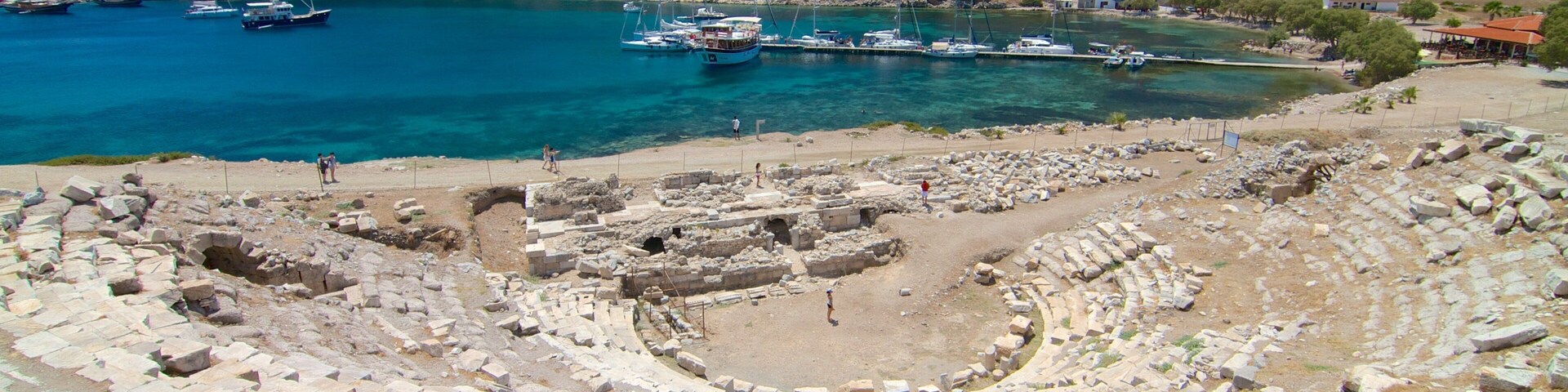 Un anfiteatro antiguo con ruinas y barcos al fondo.