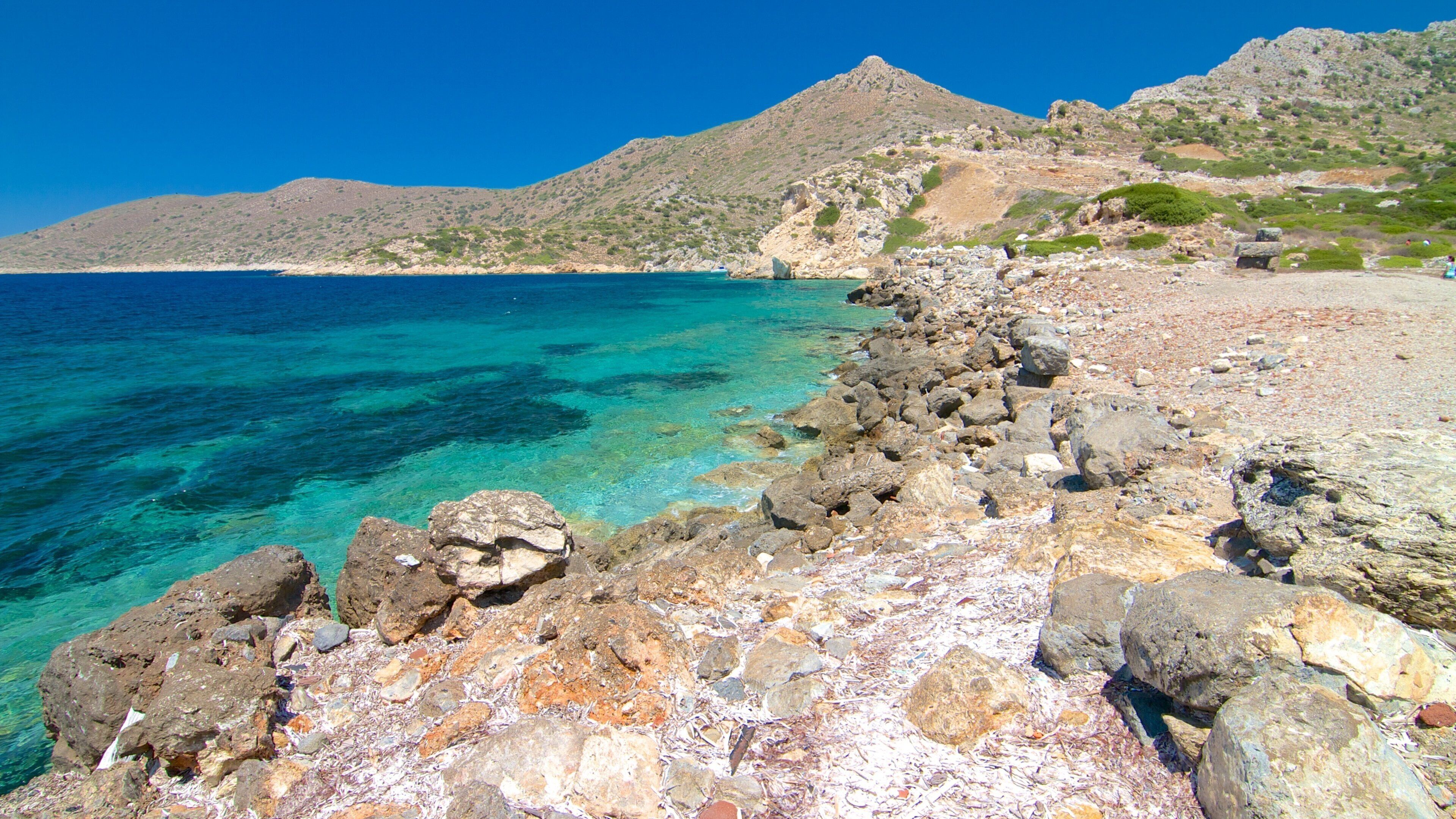 A rocky shoreline with clear turquoise water, a distant boat, and a mountainous backdrop.