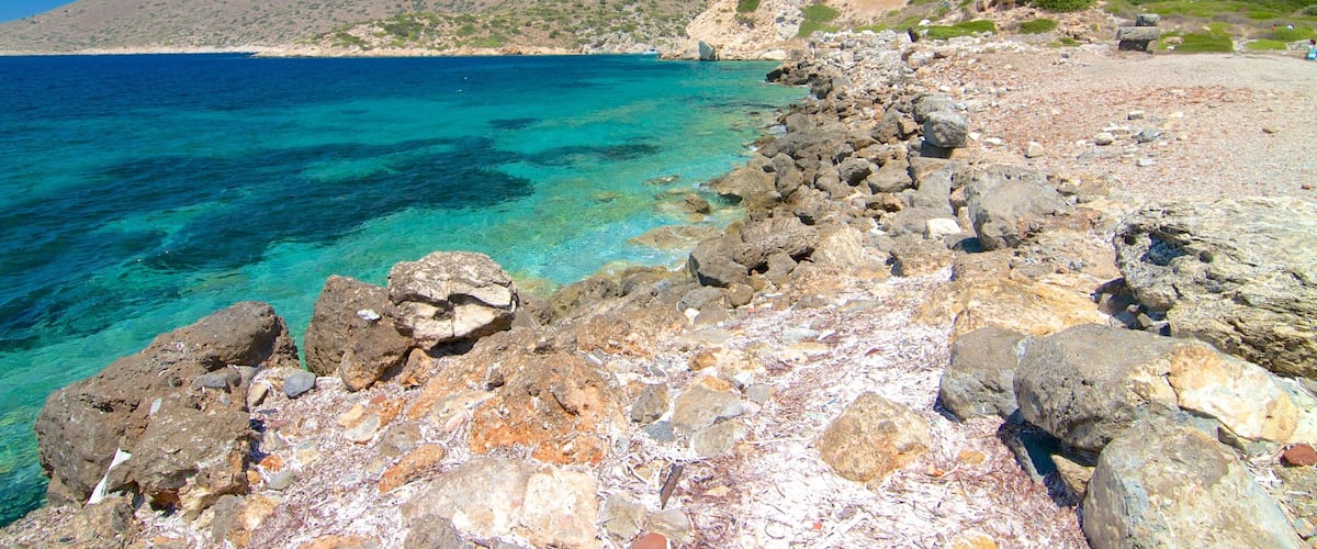 A rocky shoreline with clear turquoise water, a distant boat, and a mountainous backdrop.