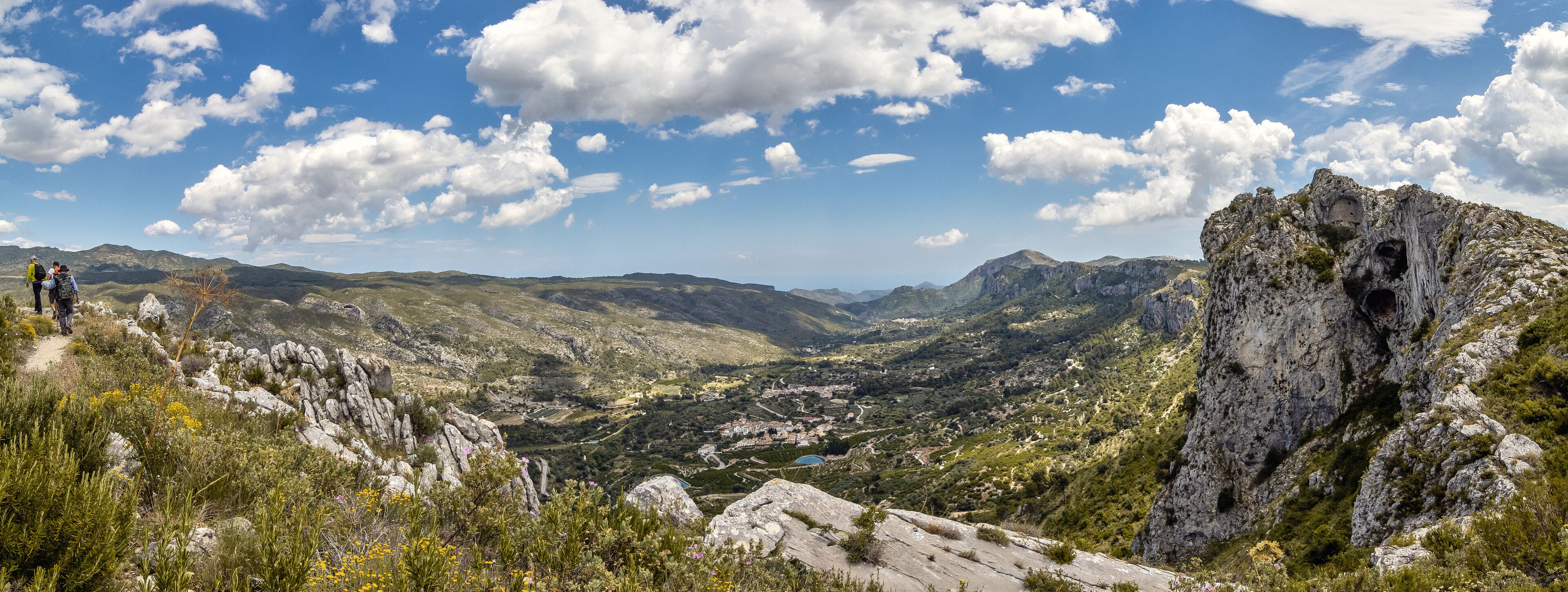 A mountainous landscape with a rocky outcrop, a valley with scattered buildings, and hikers on a trail.